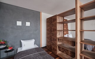 A modern bedroom with a grey wall, a wooden bookshelf, a bed with white bedding, and a small table with decorative items.
