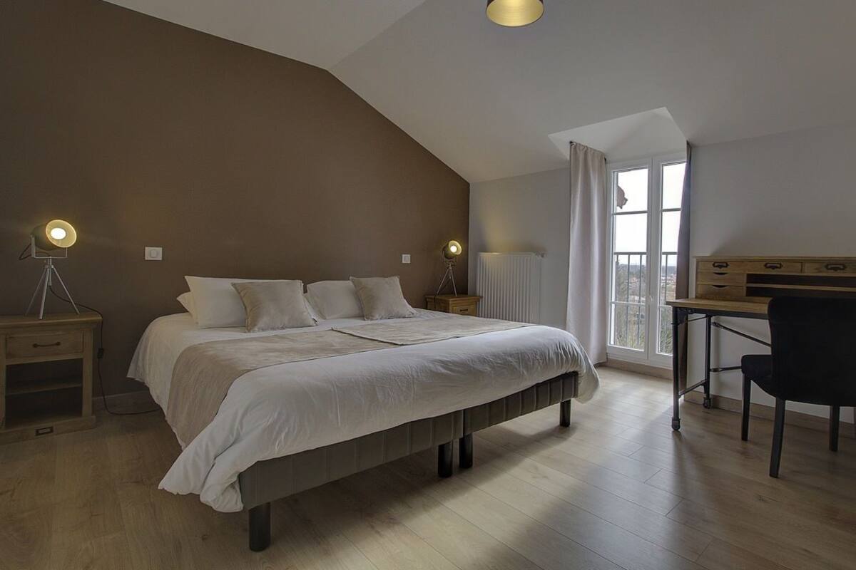 A bedroom with a large bed, bedside tables with lamps, a desk with a chair, and a view through a window.