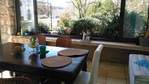 Un comedor con pared de piedra, un ventanal con vistas a zonas verdes y edificios, y una mesa dispuesta con manteles individuales y adornos.