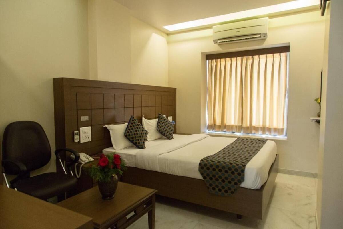 Habitación de hotel con cama, escritorio, silla, un jarrón con flores, aire acondicionado y ventana con cortinas.