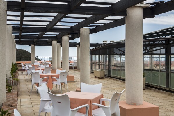An outdoor patio with white chairs and tables, covered by a pergola with concrete pillars.