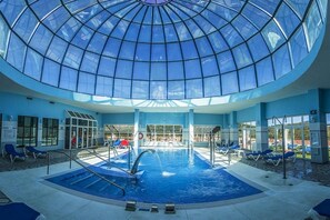 An indoor swimming pool with a glass dome ceiling, blue lounge chairs, and a clear view of the outside. - Hotel Spa Marbella Hills (Ojén)