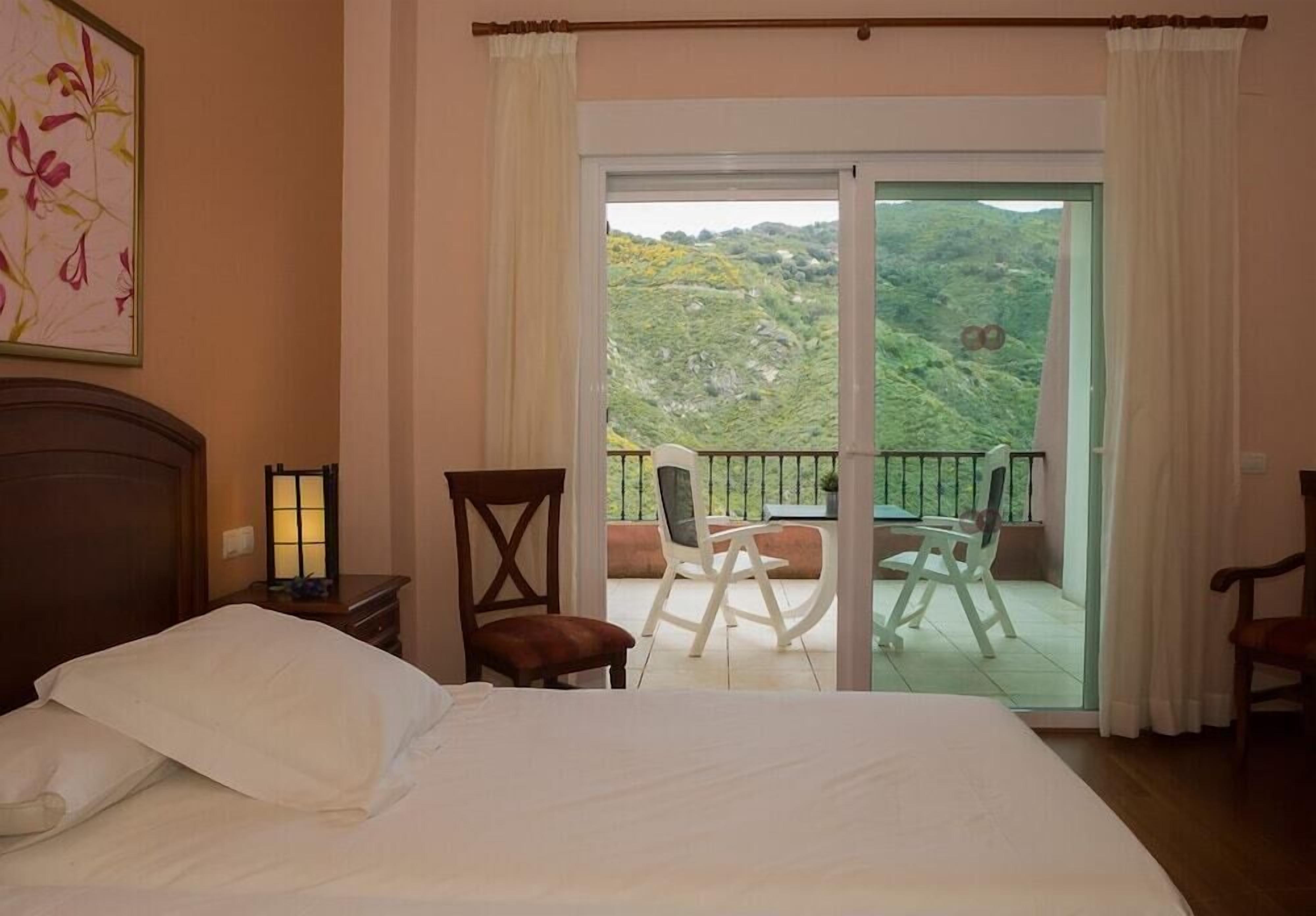 A hotel room with a bed, a nightstand with a lamp, a chair, and a balcony with a table and chairs.