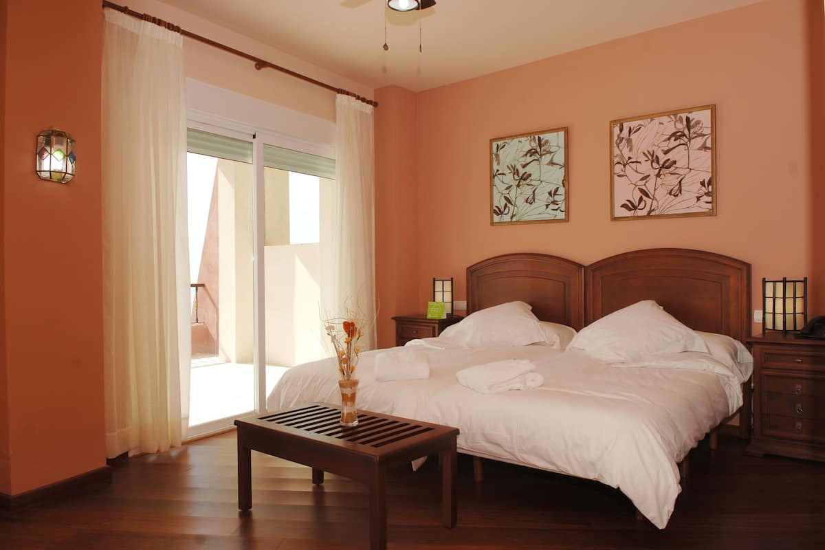 A bedroom with a bed, bedside tables, a nightstand, a window with curtains, and two wall-mounted artworks.