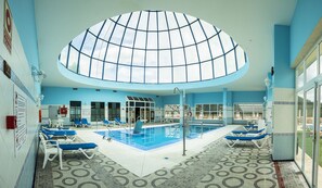Una piscina interior con techo de cristal, sillas reclinables azules y suelo de baldosas.