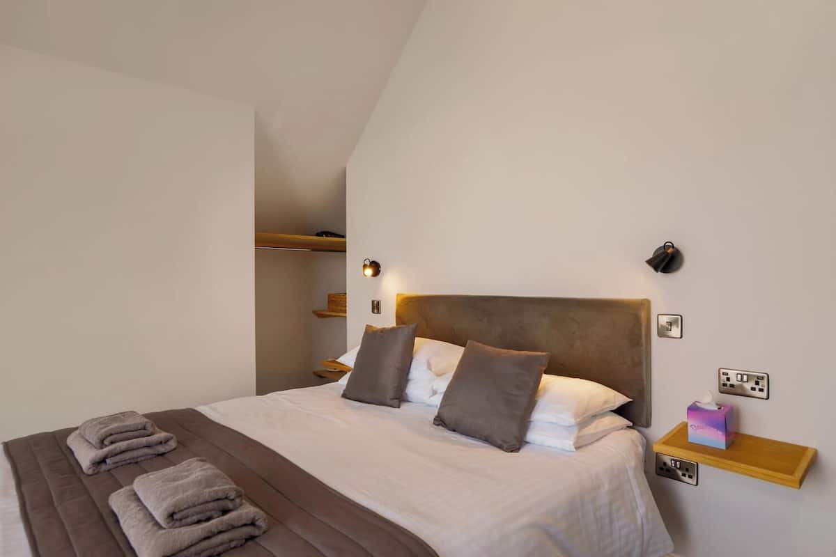 A bedroom with a bed, pillows, a bedside table, a wall-mounted light, and a shelf with a tissue box.