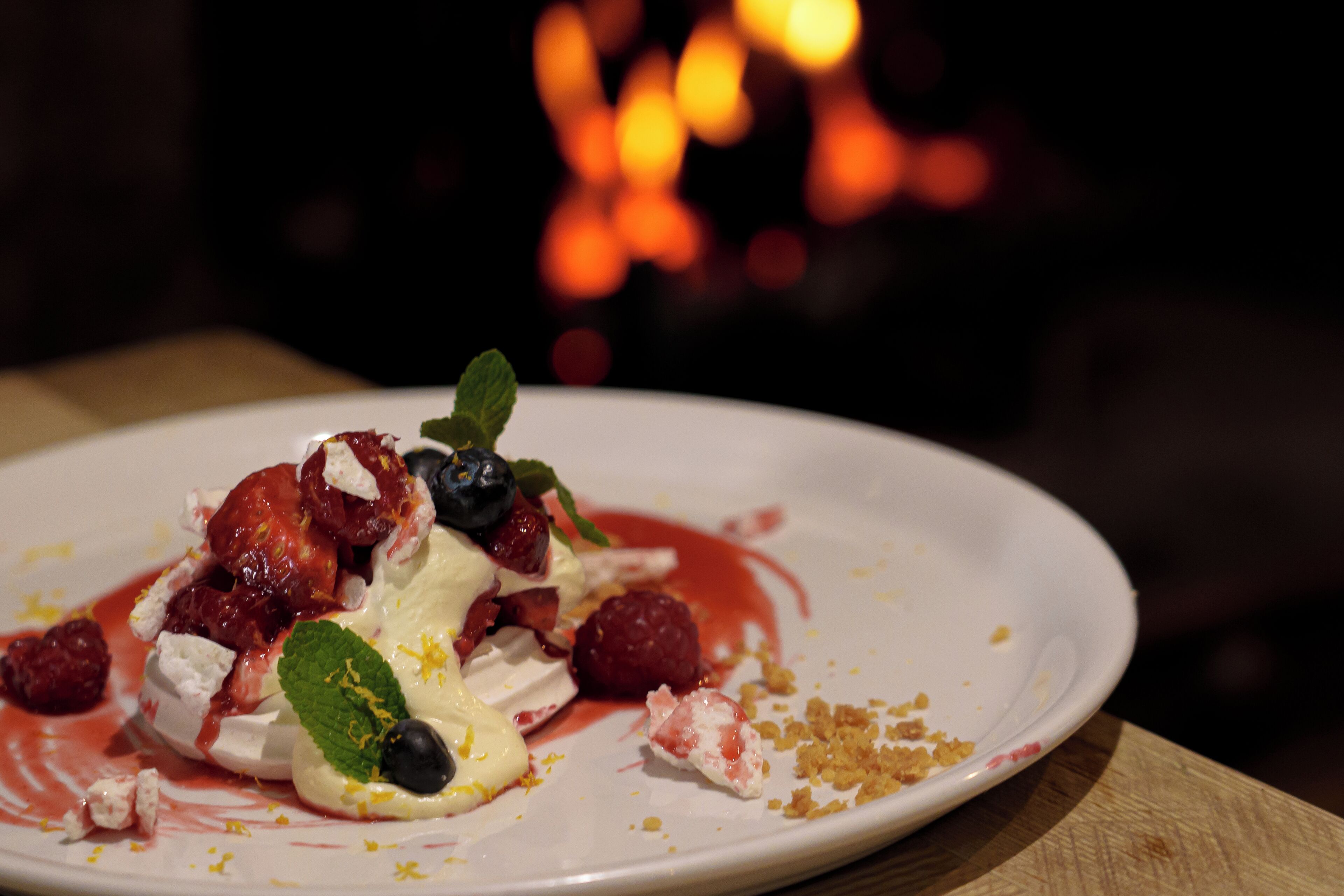 A dessert on a white plate with strawberries, blueberries, raspberries, mint, and a white cream sauce, with a blurred background of lights.