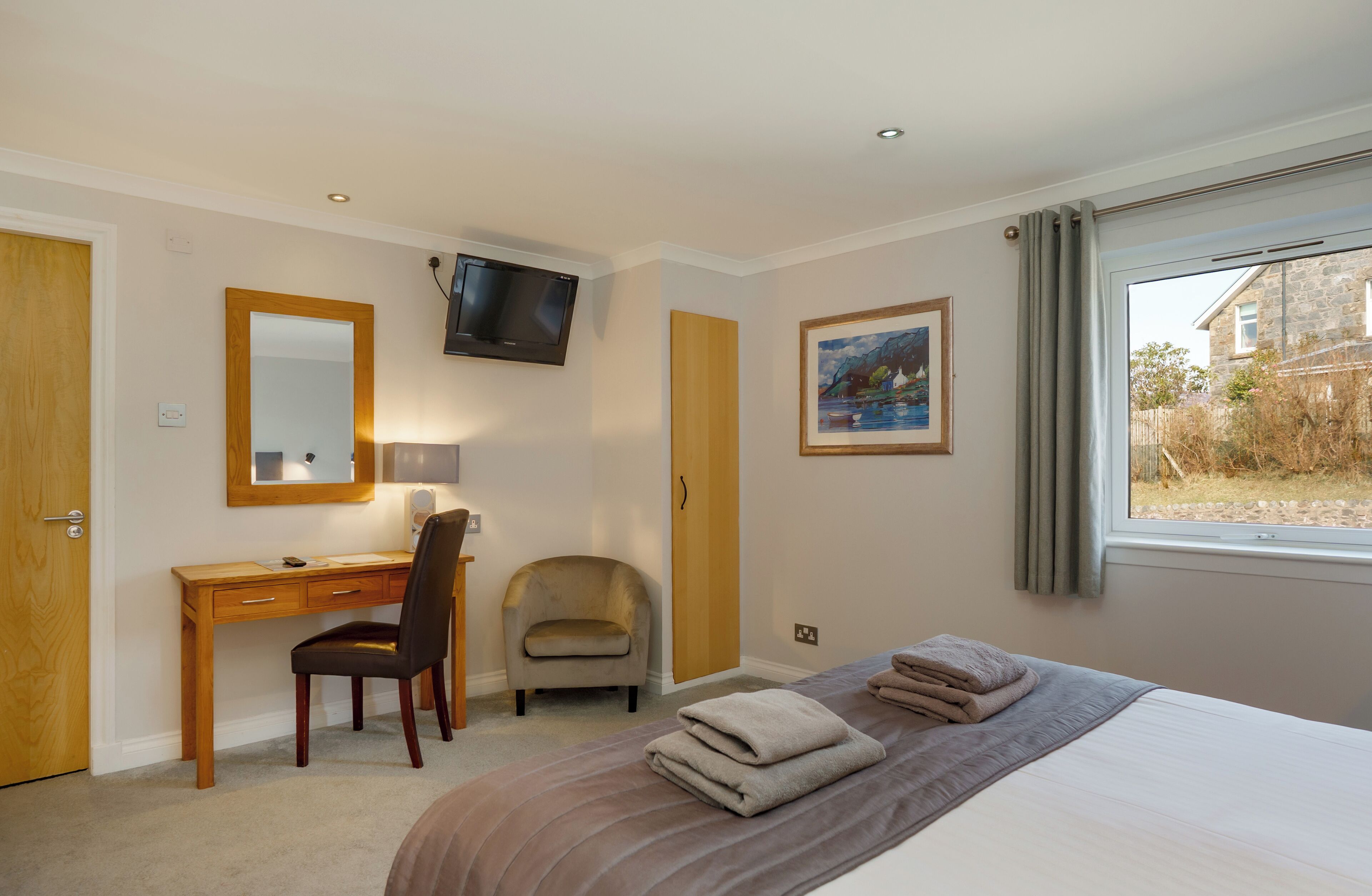A hotel room with a bed, a desk, a chair, a TV, a painting, and a window with curtains.