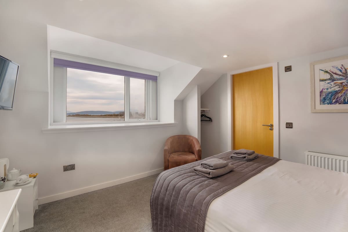 A bedroom with a bed, a window with a view, a chair, a door, and a radiator.