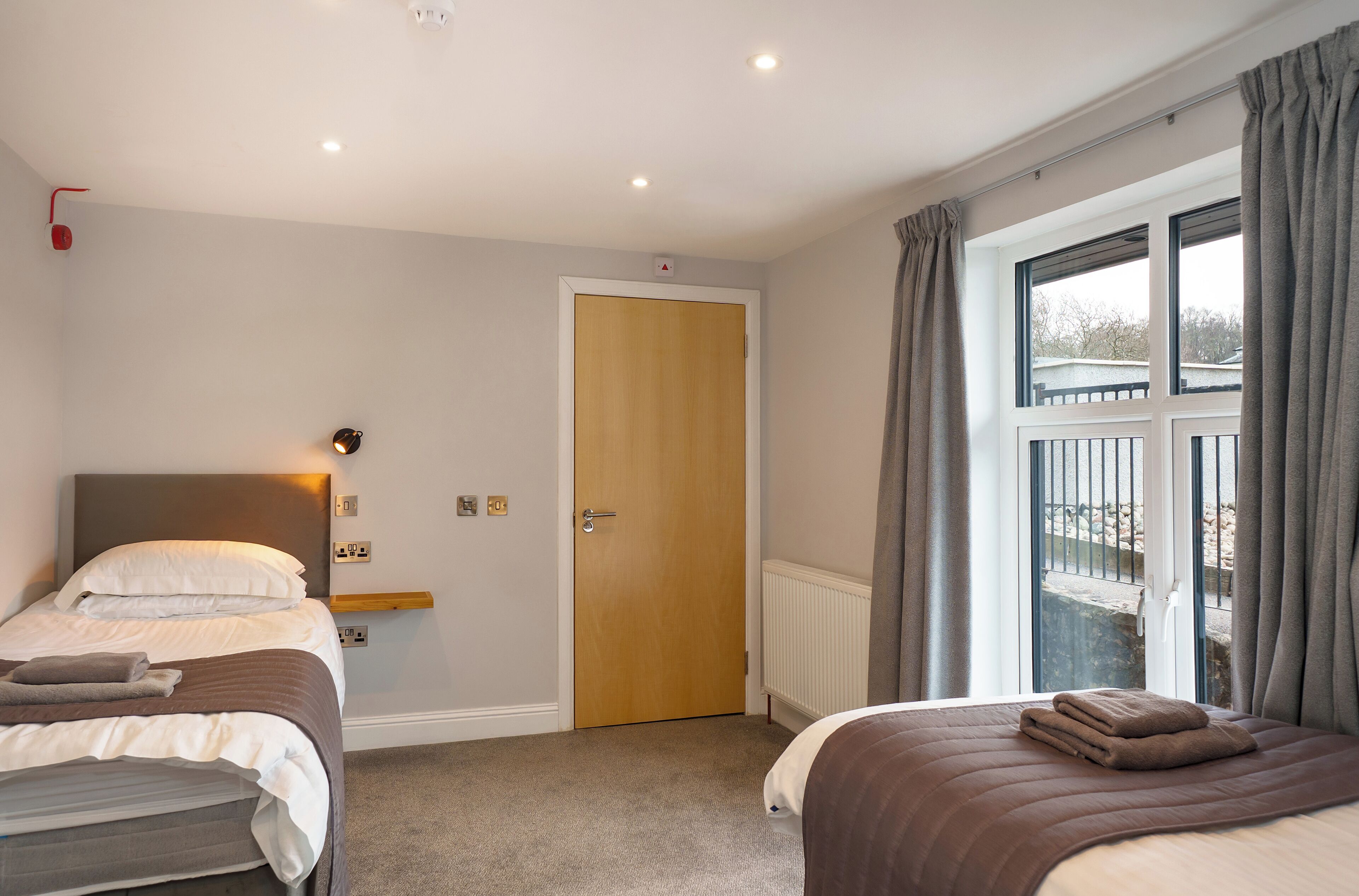 A hotel room with two beds, a wooden door, a window with curtains, and a view of the outside.