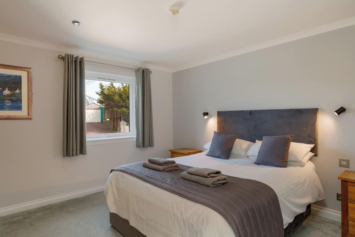 A bedroom with a large bed, a window with curtains, a bedside table, and a painting on the wall.