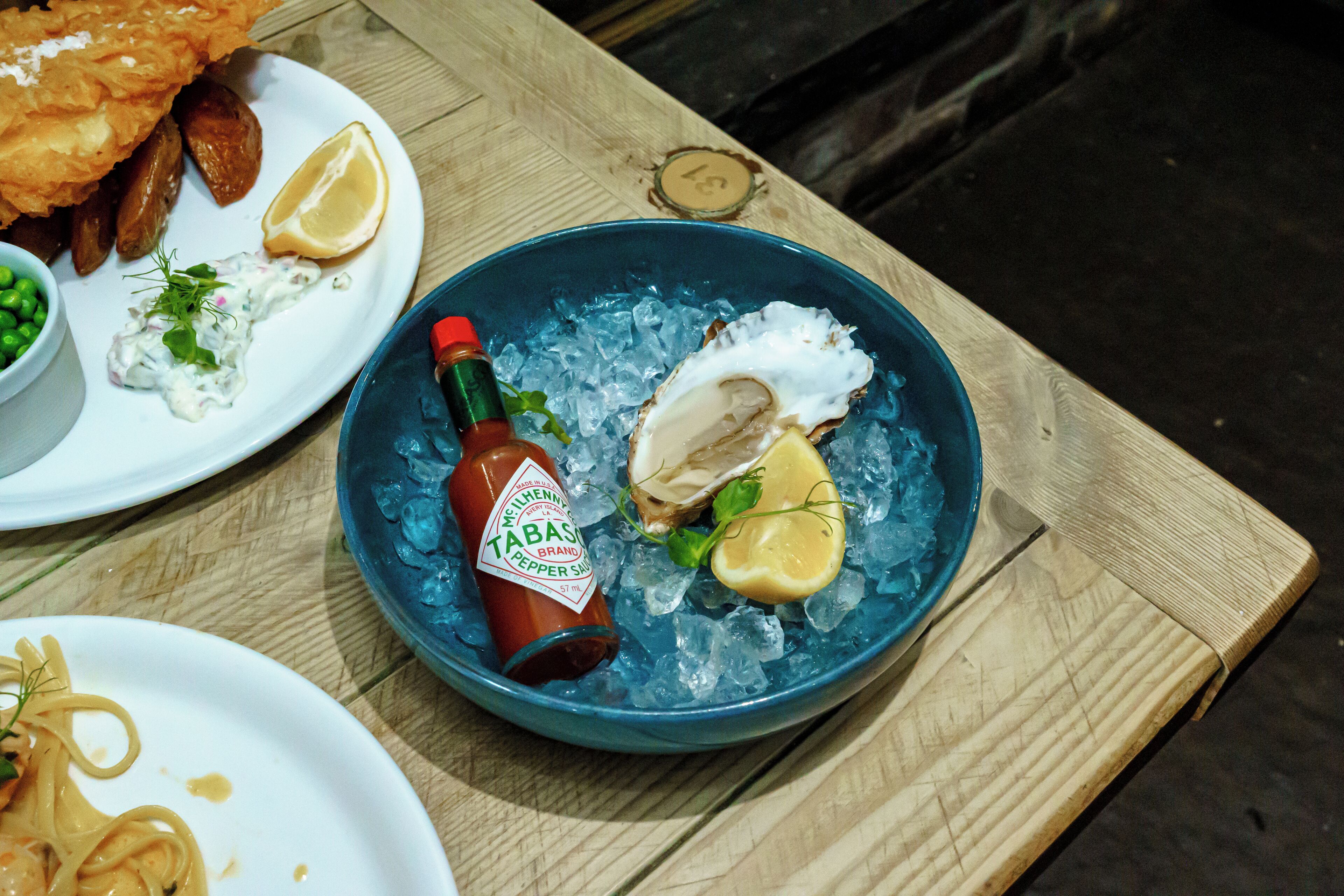 A table with a blue bowl containing an oyster, lemon, and Tabasco sauce on ice.