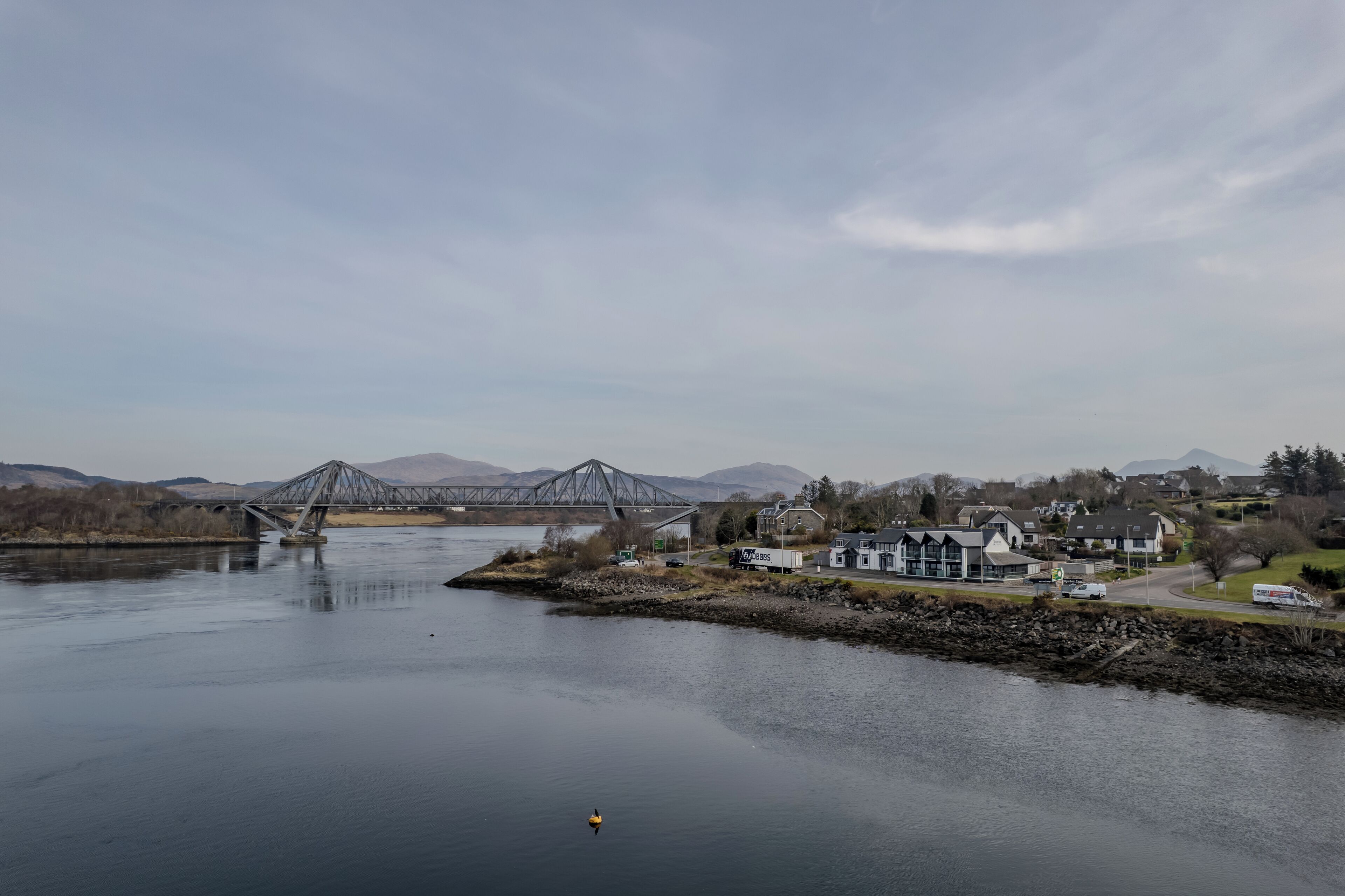 A river with a bridge, houses, and mountains in the background.
