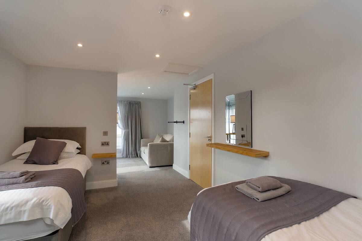 A hotel room with two beds, a sofa, a wooden shelf, and a mirror.