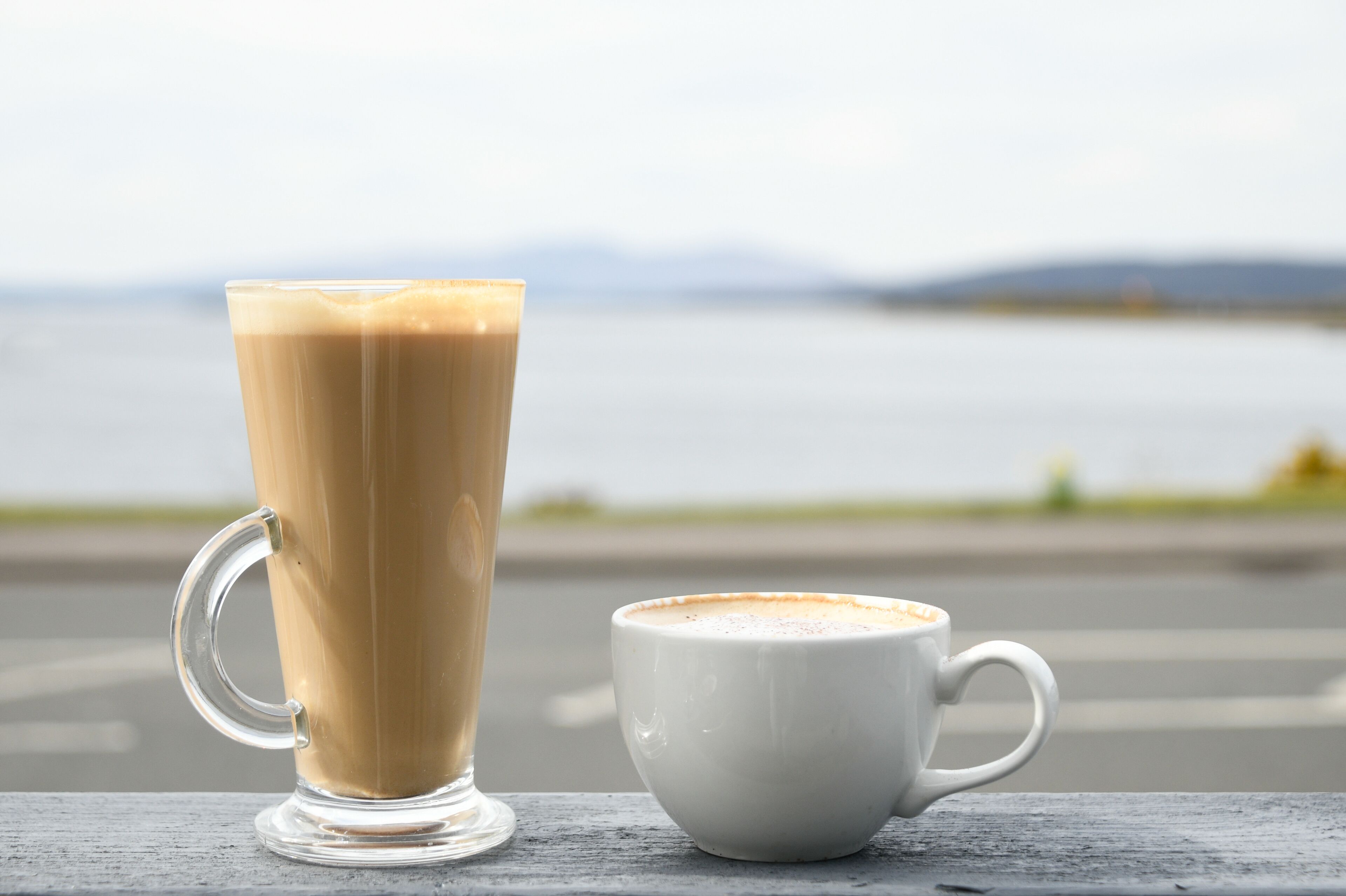 A tall glass and a short cup of coffee on a wooden surface with a view of a body of water and distant hills.