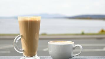 A tall glass and a short cup of coffee on a wooden surface with a view of a body of water and distant hills.