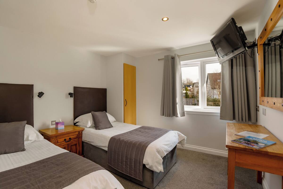 A hotel room with two beds, a wooden nightstand, a desk with a magazine, a television mounted on the wall, and a window with curtains.