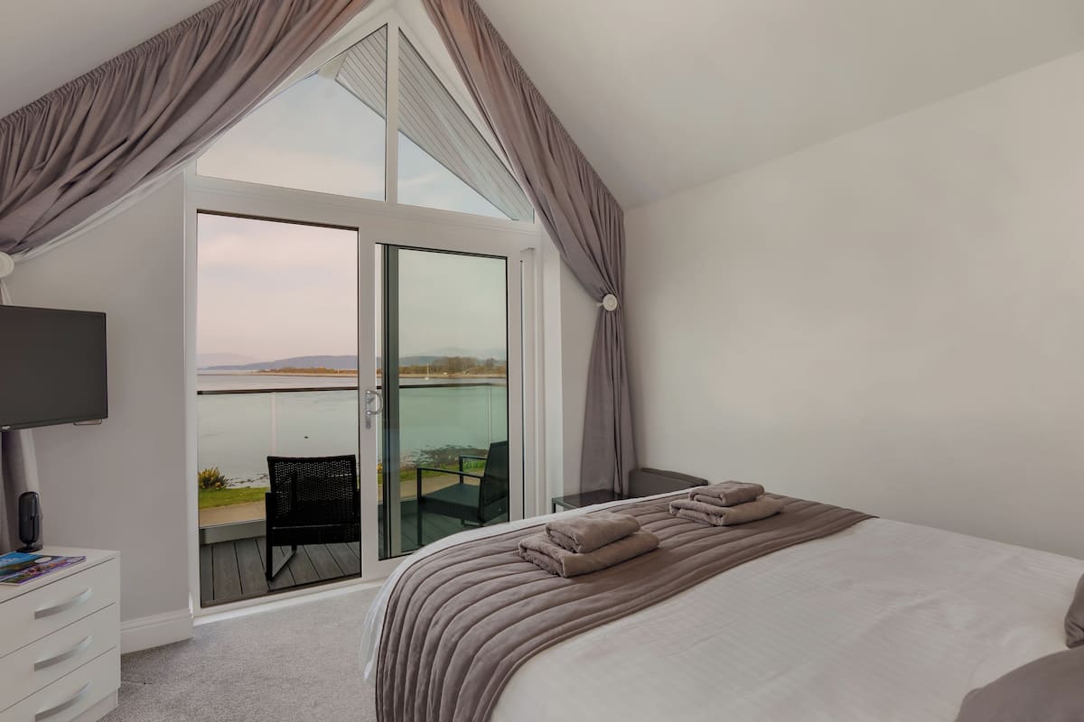 A bedroom with a large bed, a TV, a desk, and a balcony with a view of the water.