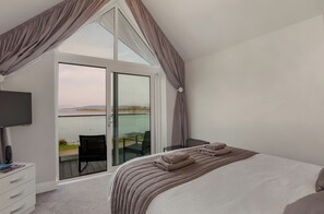 A bedroom with a large bed, a TV, a desk, and a balcony with a view of the water. - Oyster Inn (Oban)