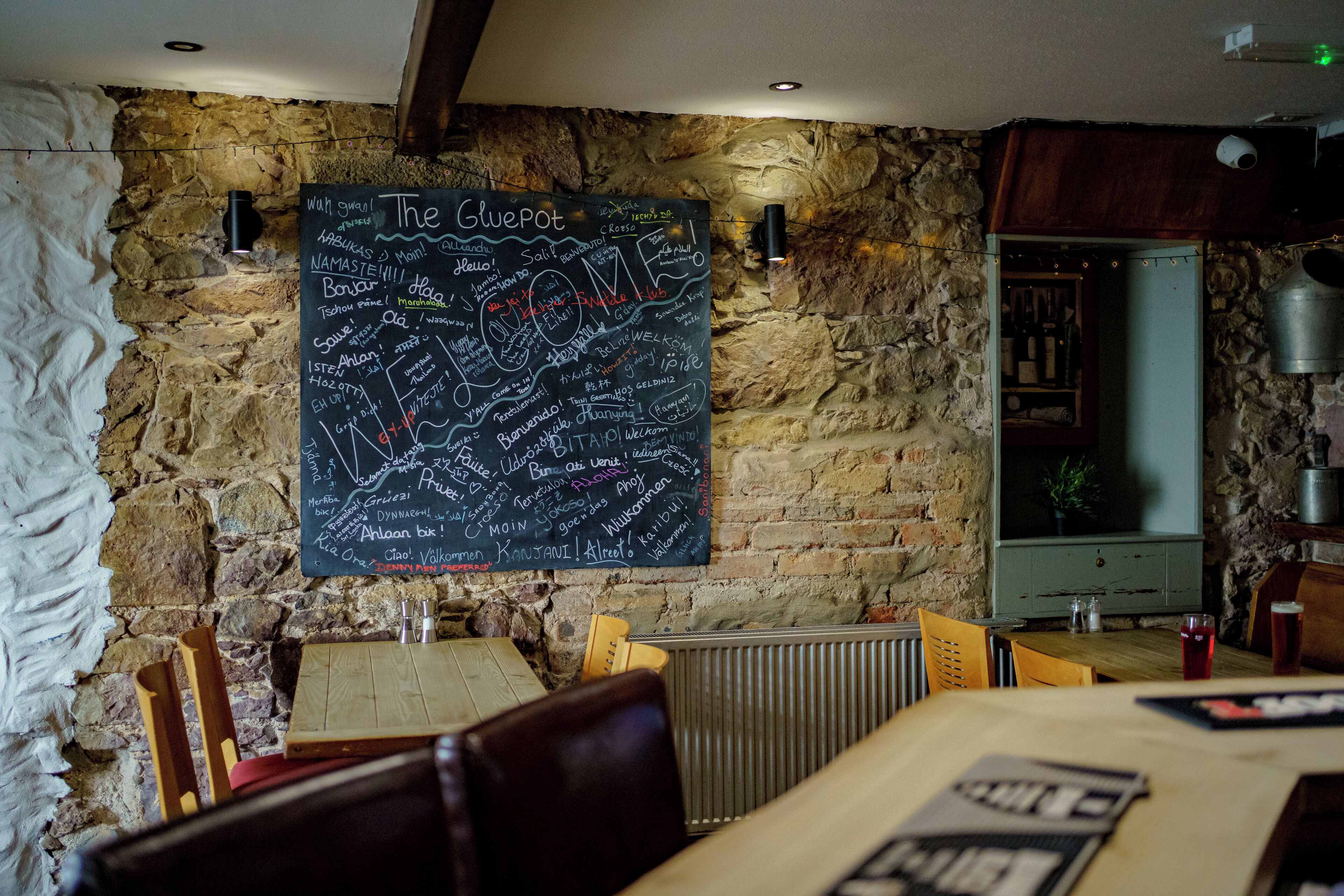 Ein Restaurantinneres mit einer Kreidetafel-Speisekarte, Steinwänden, Holzmöbeln und einem sichtbaren Küchenbereich.