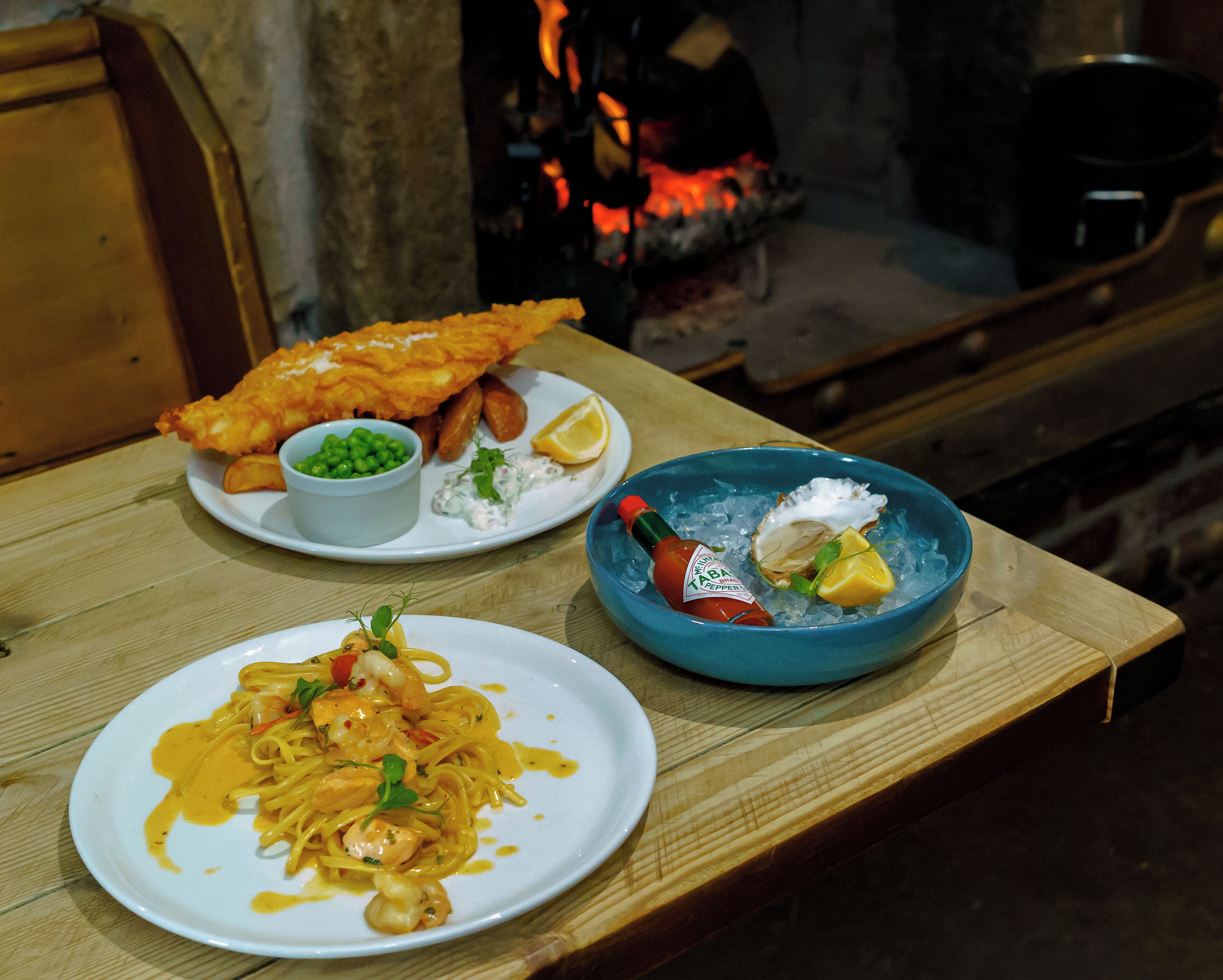 A wooden table with three plates of food: fish and chips, pasta with shrimp, and a seafood dish with lemon and herbs.