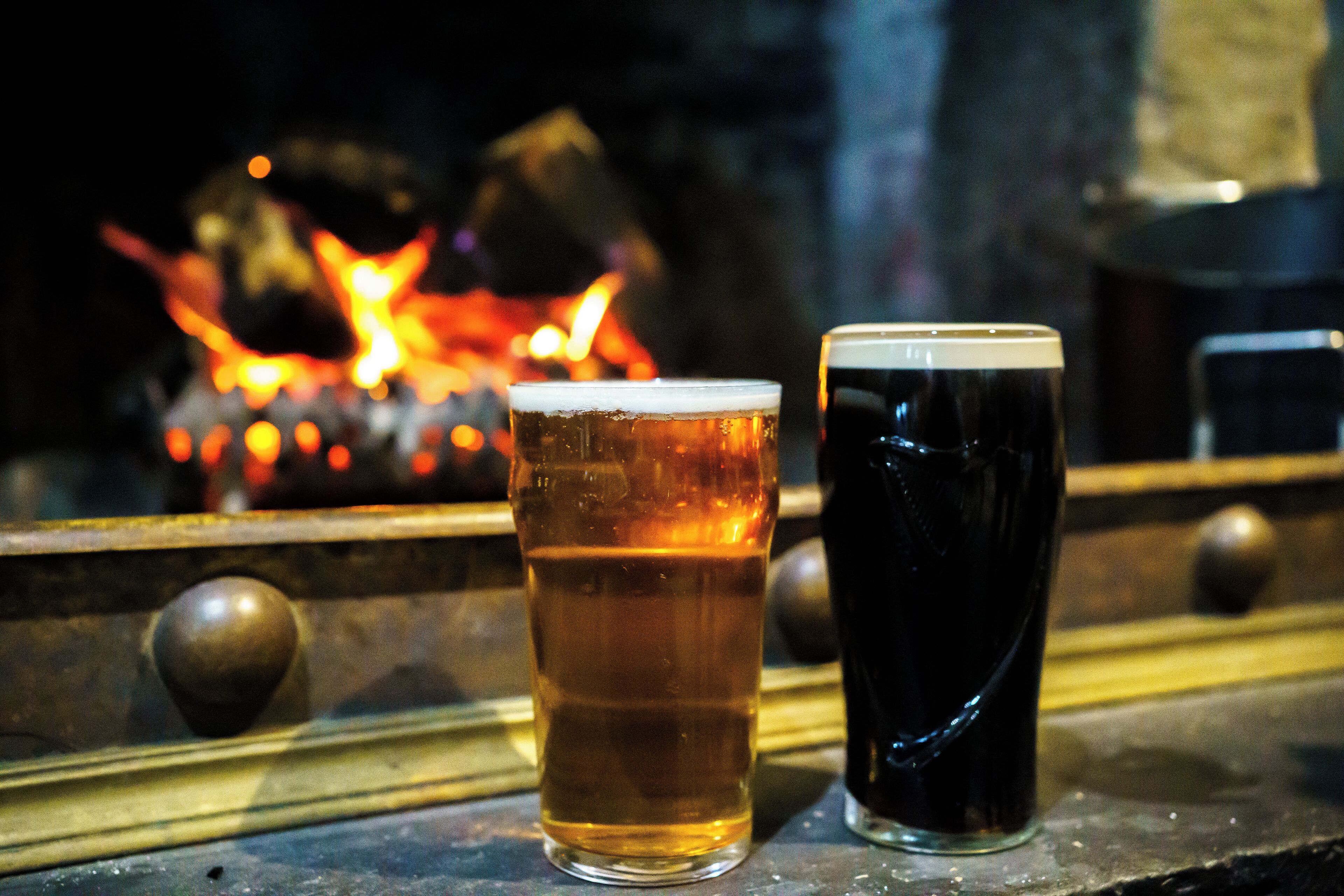 Two glasses of beer on a bar counter with a fire in the background.