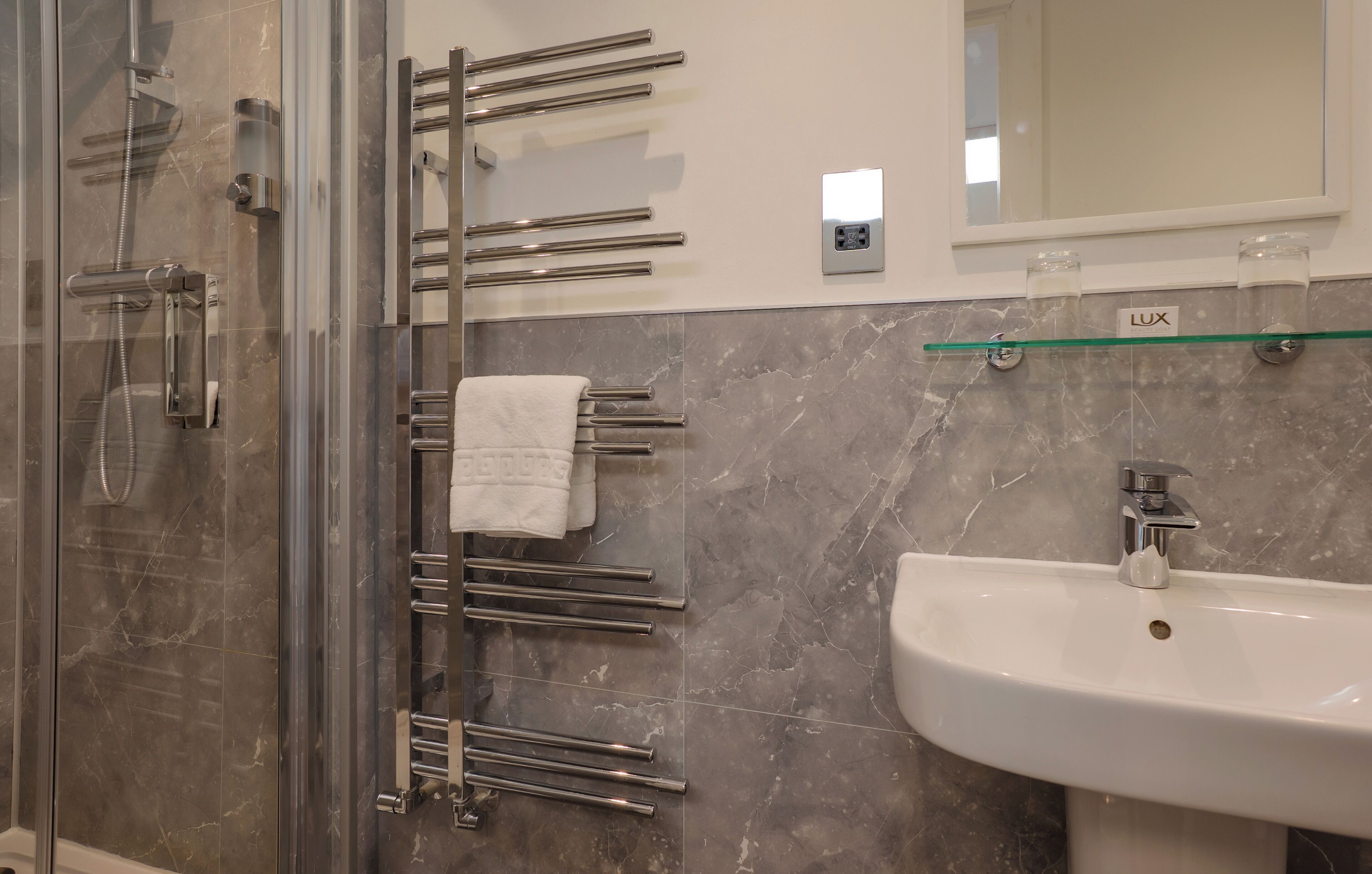 Et moderne badeværelse med en glasbrusekabine, en glashylde med toiletartikler, en hvid vask og et håndklædetørrestativ.
