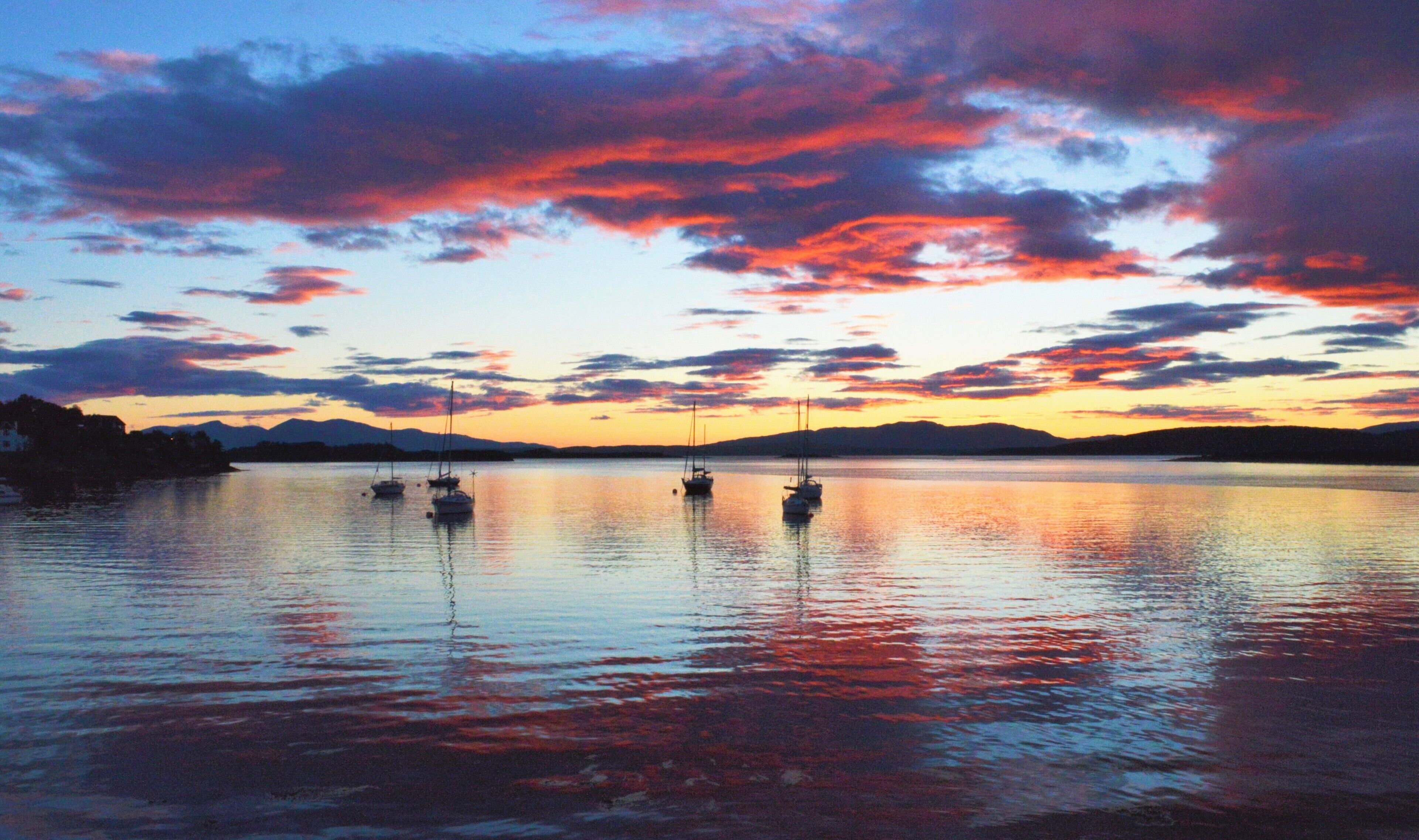 A calm body of water with several boats anchored, surrounded by a mountainous landscape under a colorful sunset sky.