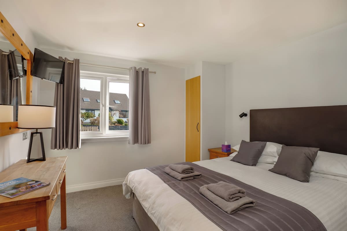 A hotel room with a large bed, a desk with a lamp, a TV mounted on the wall, and a window with curtains.