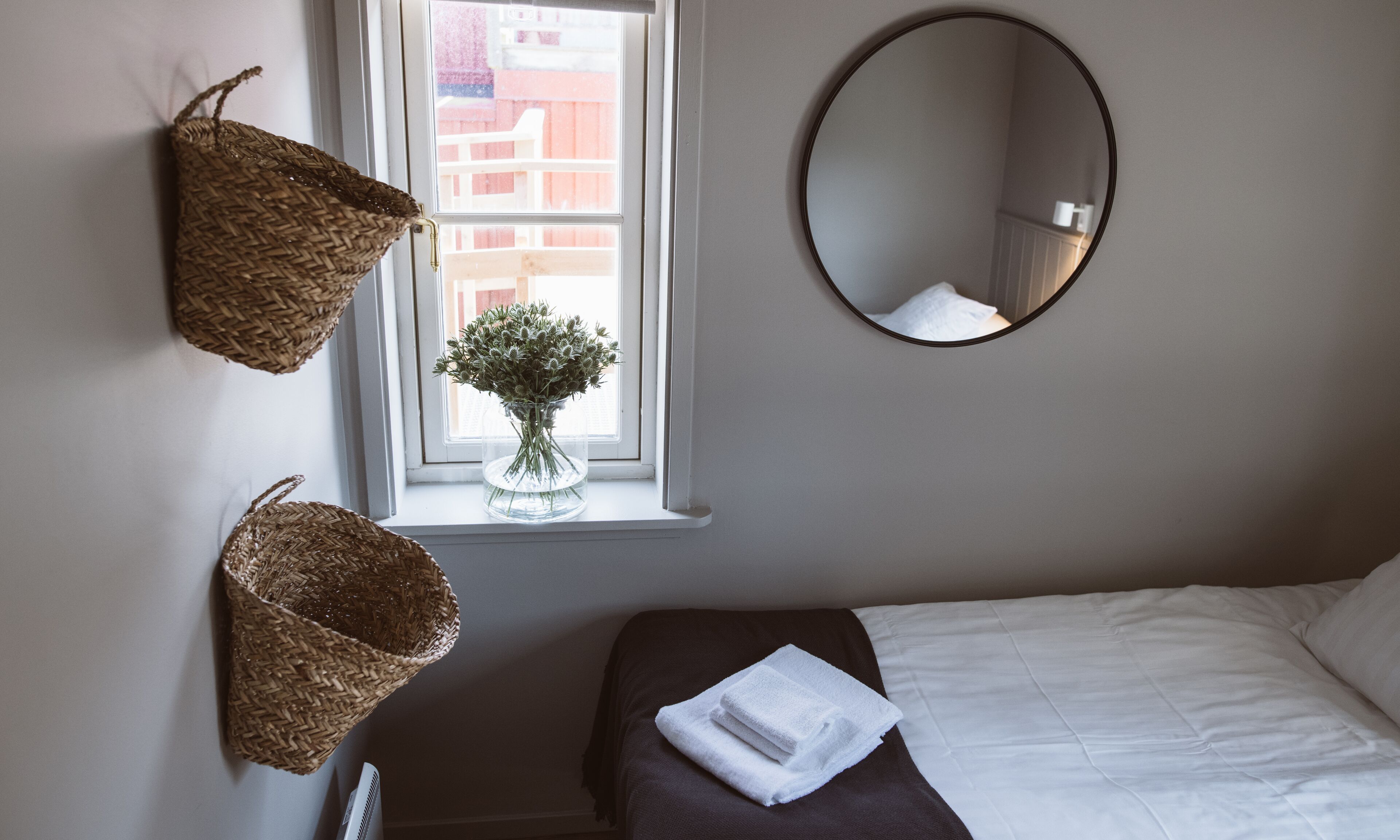Quarto com cama, janela com vista para edifícios, espelho e duas cestas de palha.