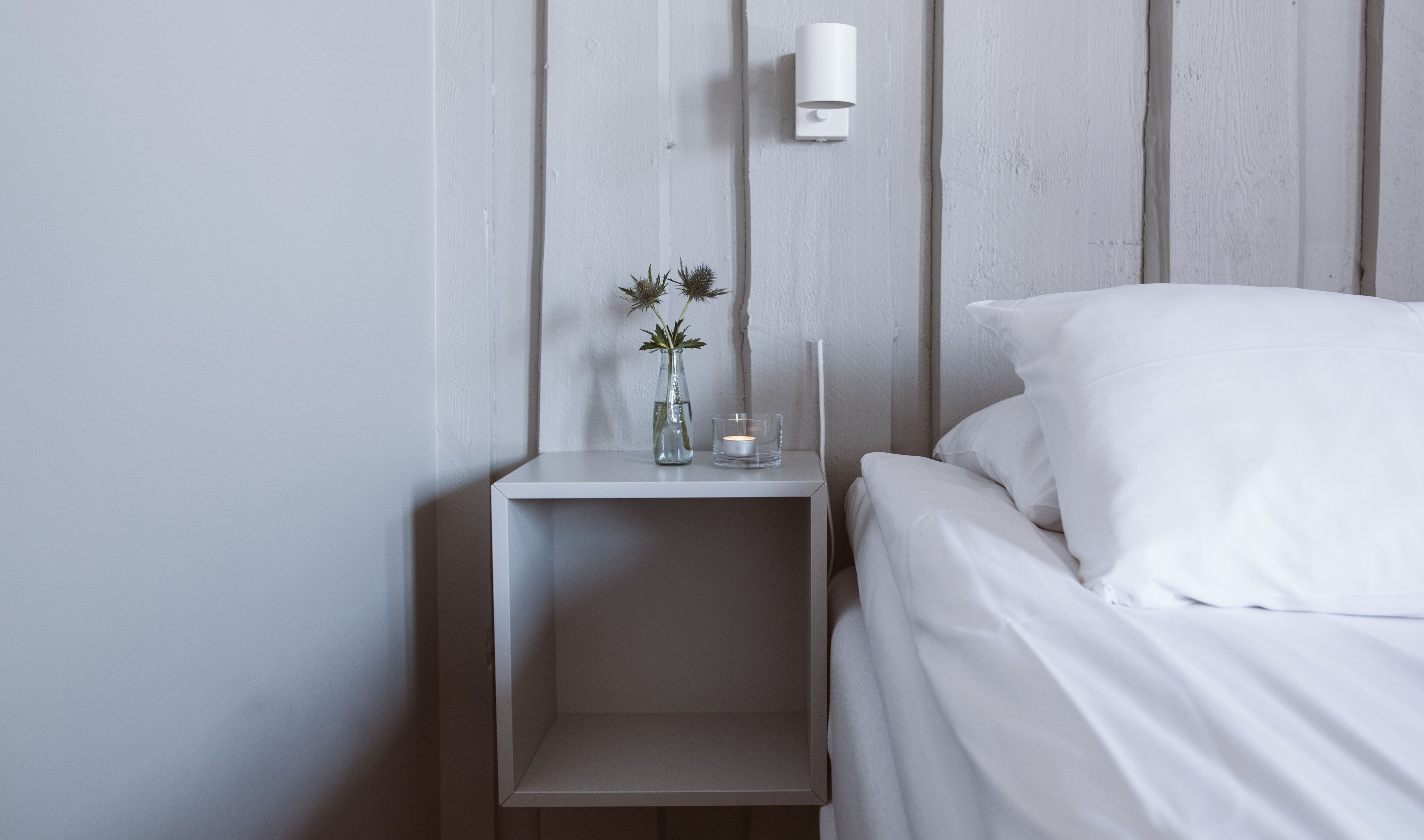 A minimalist bedroom with a white bed, a small bedside table, and a wall-mounted light.