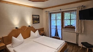 Ein Doppelbett mit Kopfteilen, eine Deckenleuchte, ein Fenster mit Vorhängen, ein Heizkörper und ein an der Wand befestigtes Fernsehgerät.