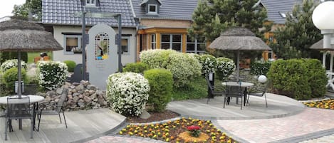 An outdoor patio area with round tables and chairs, surrounded by flower beds and a building with a grey tiled roof.