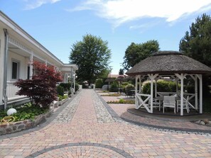 A paved pathway leading to a gazebo with outdoor seating, surrounded by greenery and a building with a white facade. - Haffblick (1) - Landhaus Mönkebude (Mönkebude)