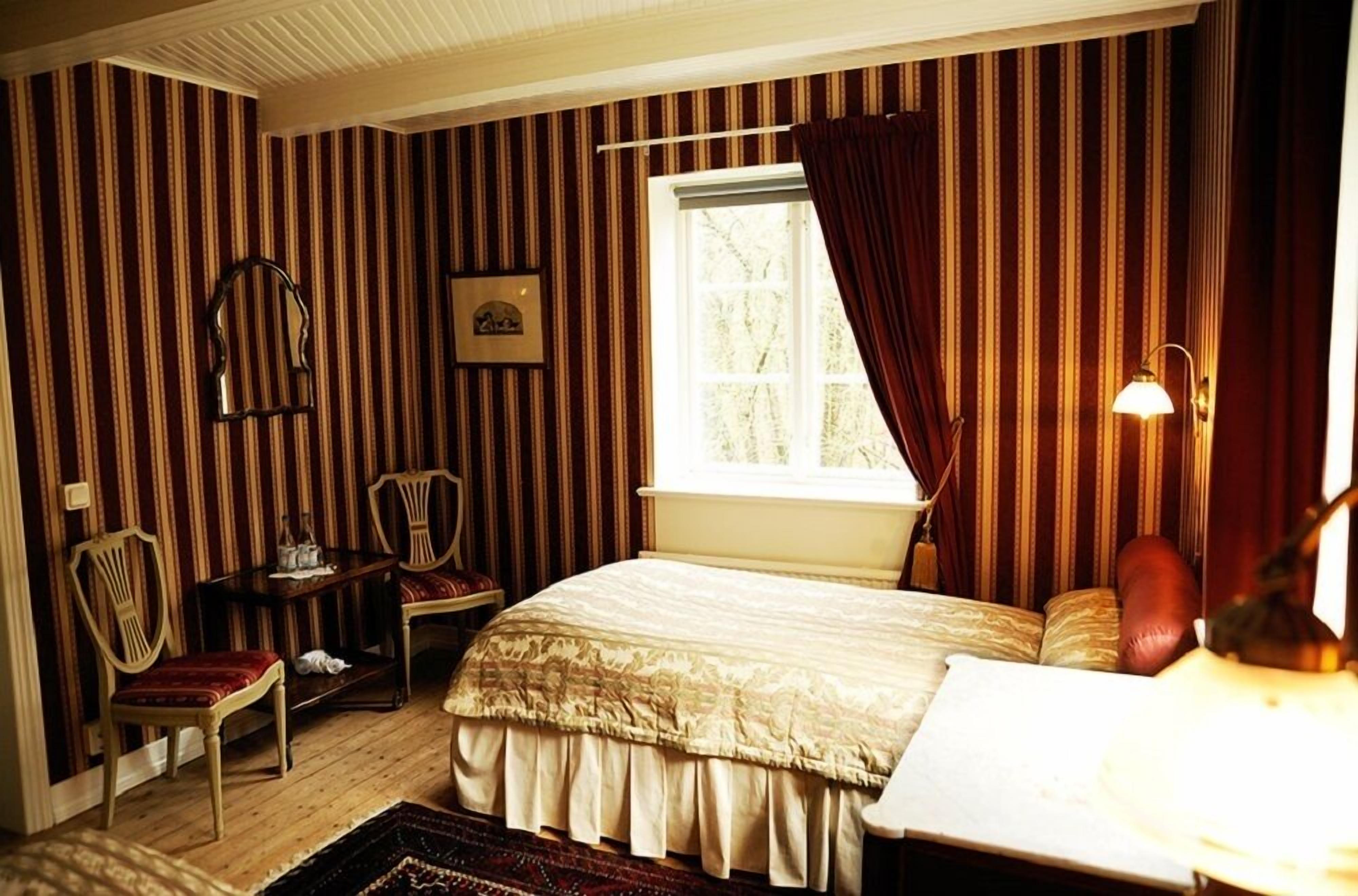 A room with striped wallpaper, a bed with a patterned bedspread, a chair, a desk, a window with curtains, and a lamp.