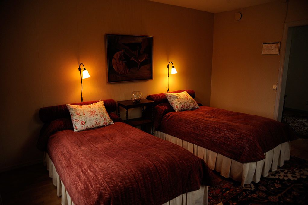 Two single beds with red bedding and white curtains, a small table with a glass, and a painting on the wall.