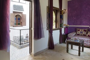 Ein Zimmer mit Balkon, einem Bett mit gemusterten Betttextilien, einem kleinen Tisch mit Dekoration und einer violetten Wand.