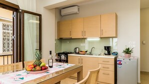 A compact kitchen with wooden cabinets, a dining table with chairs, a refrigerator, and a window with a decorative screen. - Alektor Studios & Apartments (Zakynthos)
