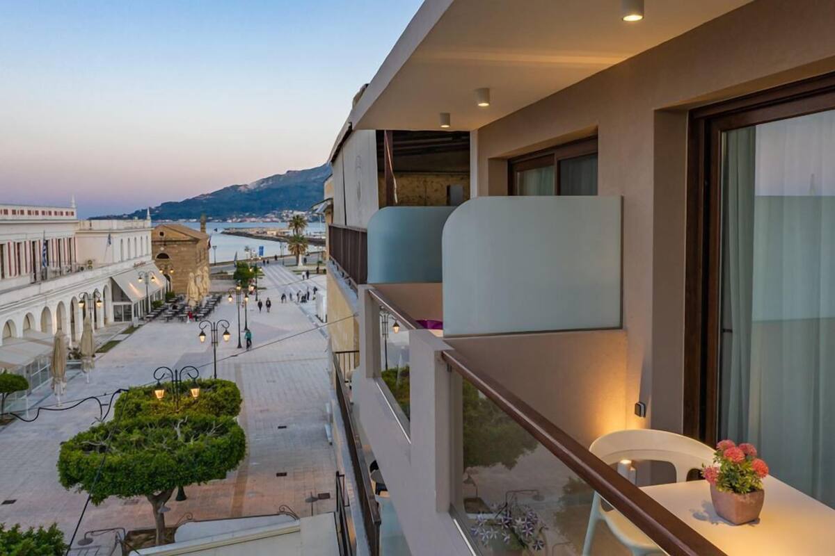 A balcony with a potted plant, overlooking a coastal promenade with buildings and a view of the sea.