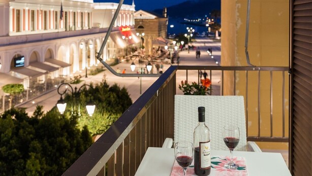 A balcony with a table set for two, a bottle of wine, and glasses. The view includes a street with buildings and a mountain in the background. - Alektor Studios & Apartments (Zakynthos)