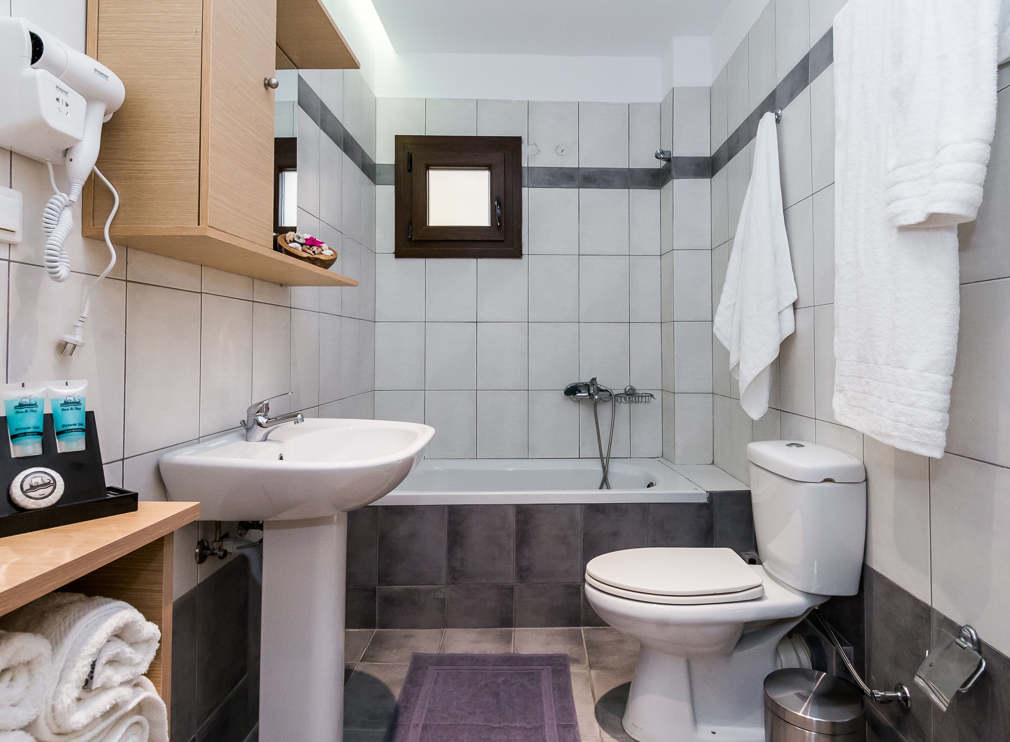Une salle de bain moderne équipée d’un lavabo, de toilettes et d’une baignoire. On y trouve un sèche-cheveux fixé au mur, un porte-serviettes et une petite étagère contenant des articles de toilette.