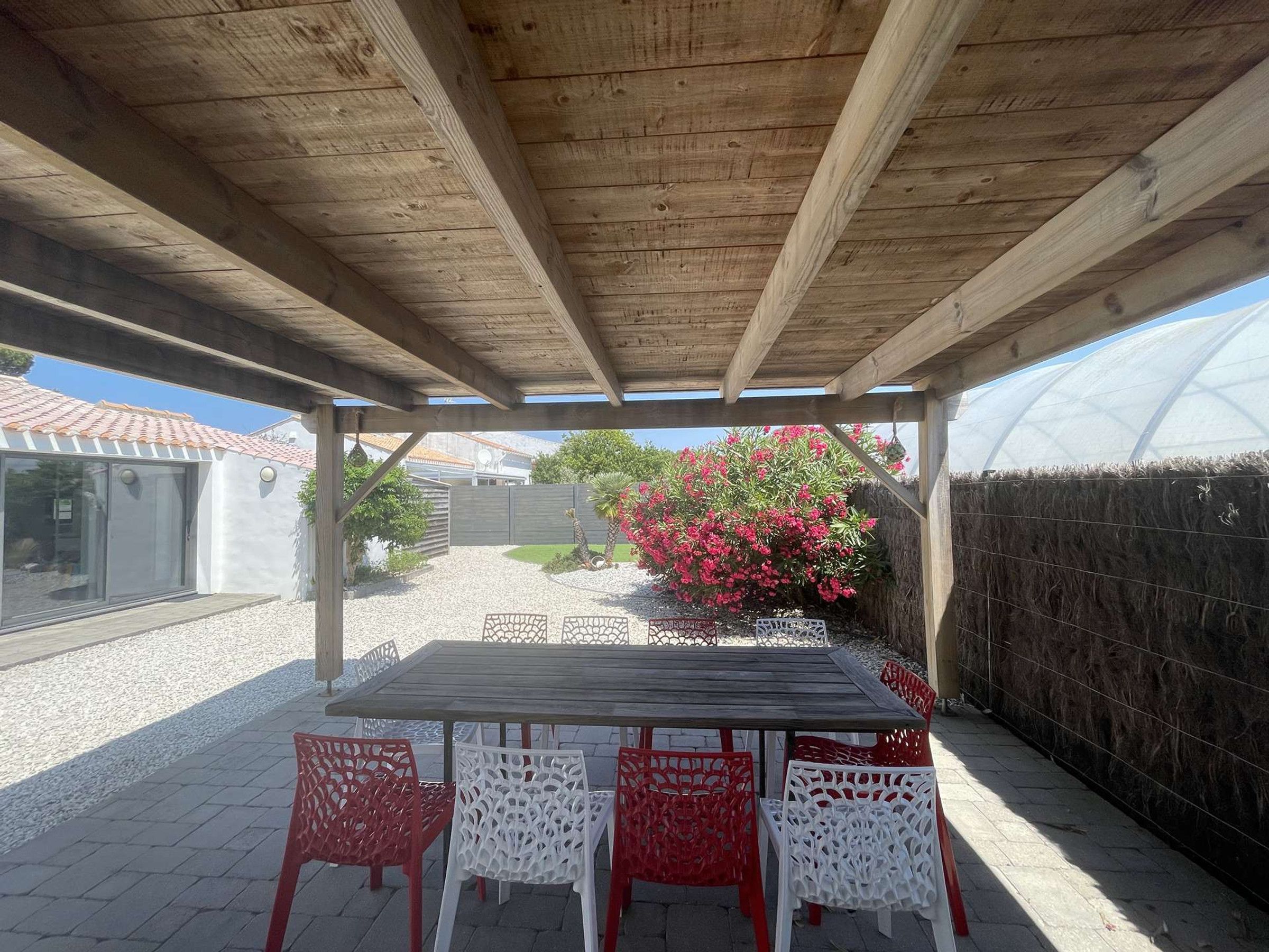 An outdoor patio with a wooden pergola, a table with chairs, and a garden with red flowers.