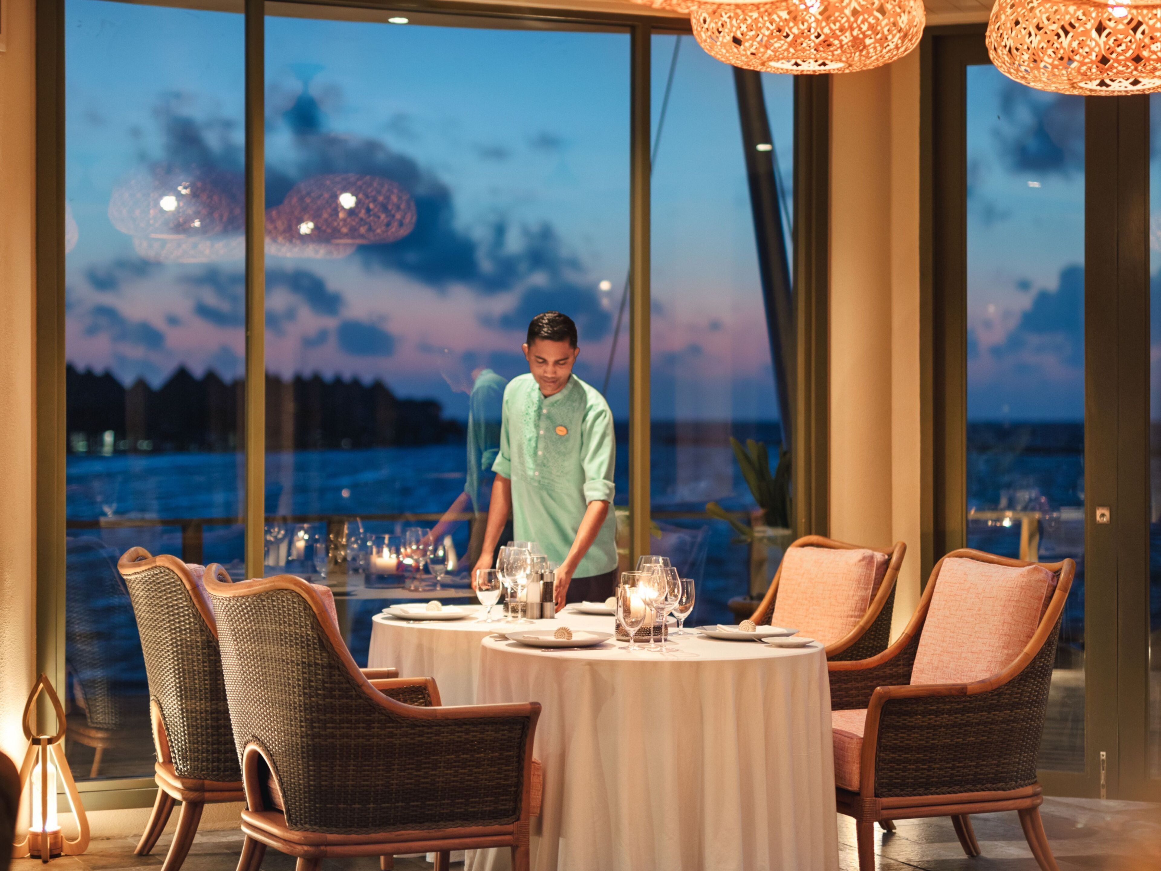 A dining area with a table set for four, large windows, wicker chairs, and a view of the sea.
