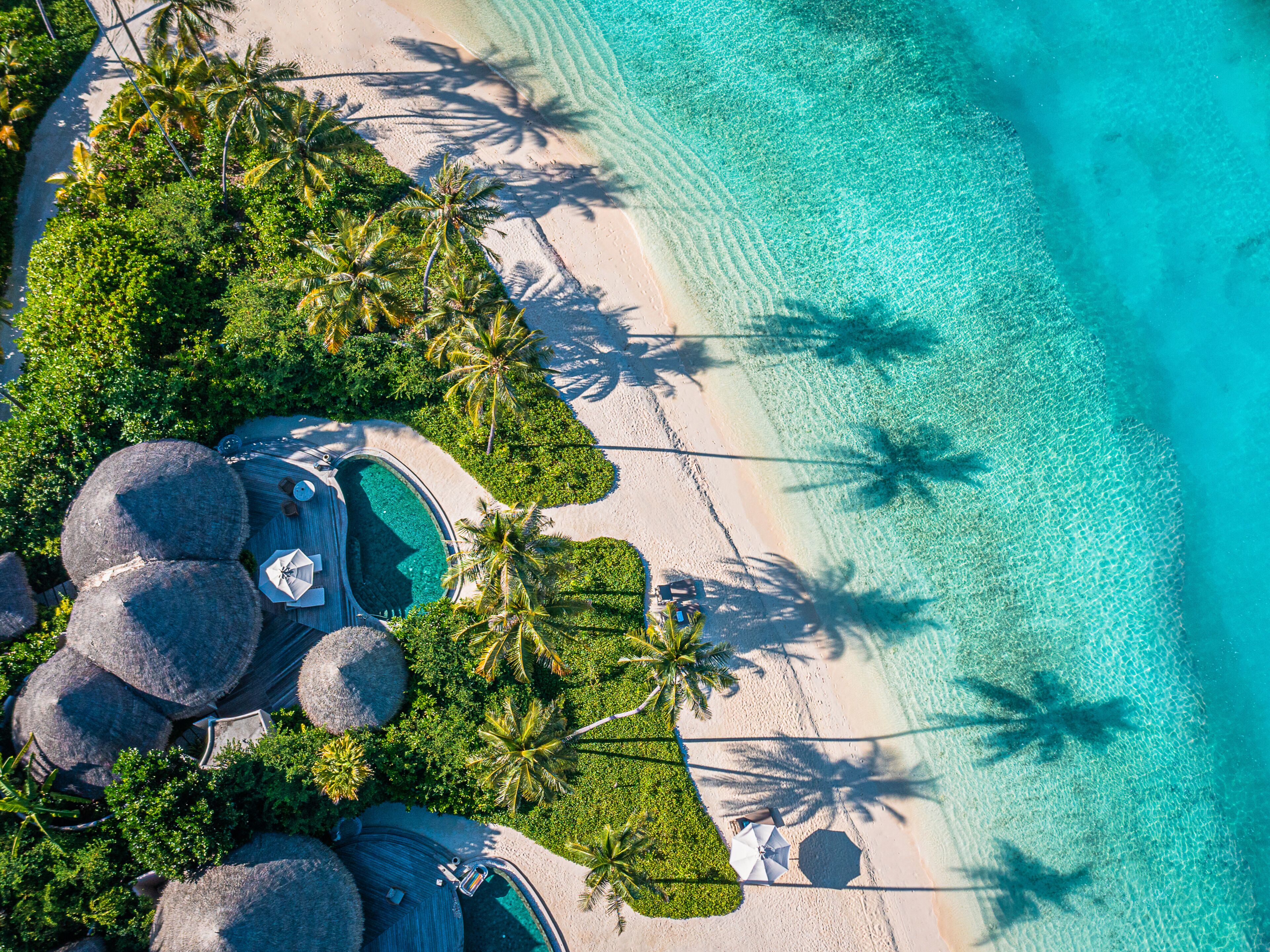 Vista aérea de un resort con un mar turquesa cristalino, palmeras y una piscina rodeada de exuberante vegetación.