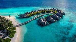 Un complejo de bungalows sobre el agua, con un puente de acceso, aguas turquesas cristalinas y una playa de arena.