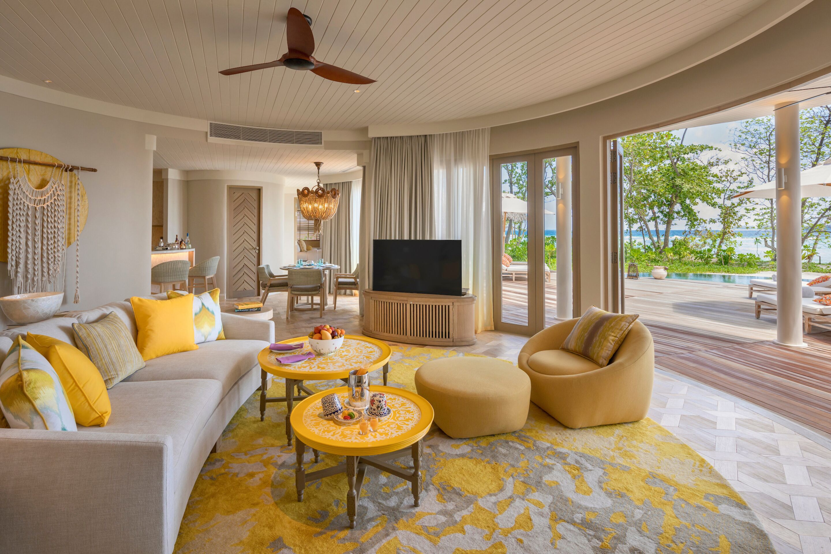A spacious living room with a large TV, yellow and white furniture, and a view of the ocean.