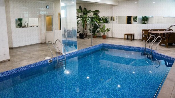 A swimming pool with clear blue water, surrounded by white tiled walls and a tiled floor. There are potted plants and a wooden bench near the pool. - Sapsan (Pavlodar)