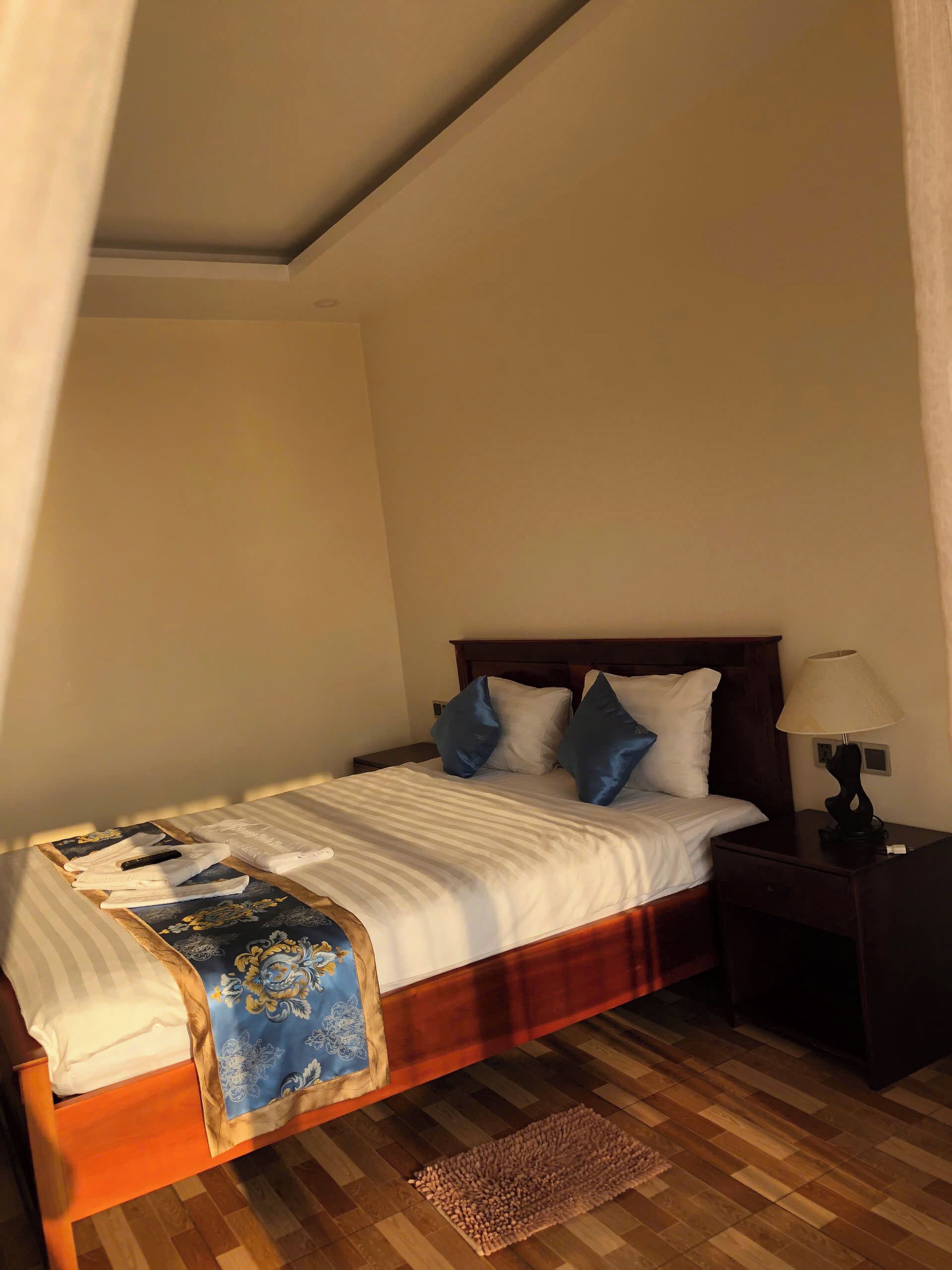 Ein ordentlich gemachtes Bett mit einer blau-weißen, gemusterten Decke, einem hölzernen Kopfteil und einem Nachttisch mit Lampe.