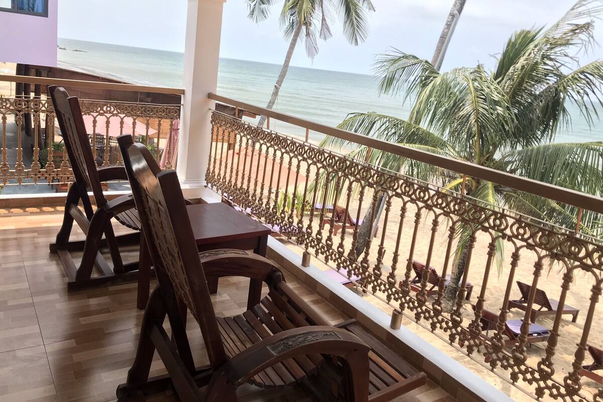 Un balcón con sillas y mesa de madera, con vista a una playa con palmeras y el océano.