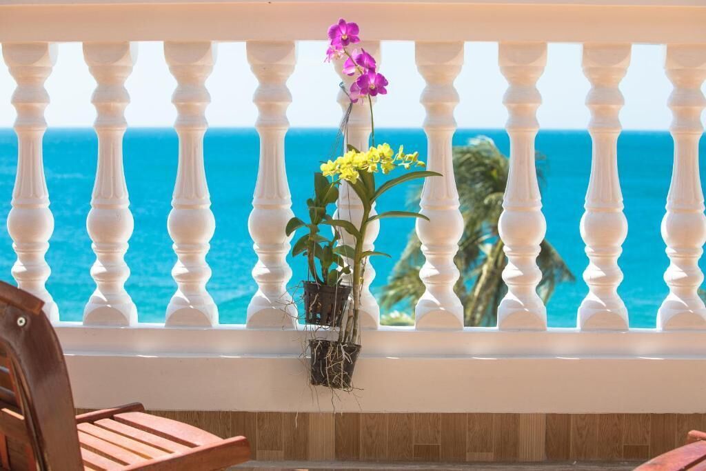 Uma varanda com balaustrada branca, um vaso com flores e vista para o oceano.